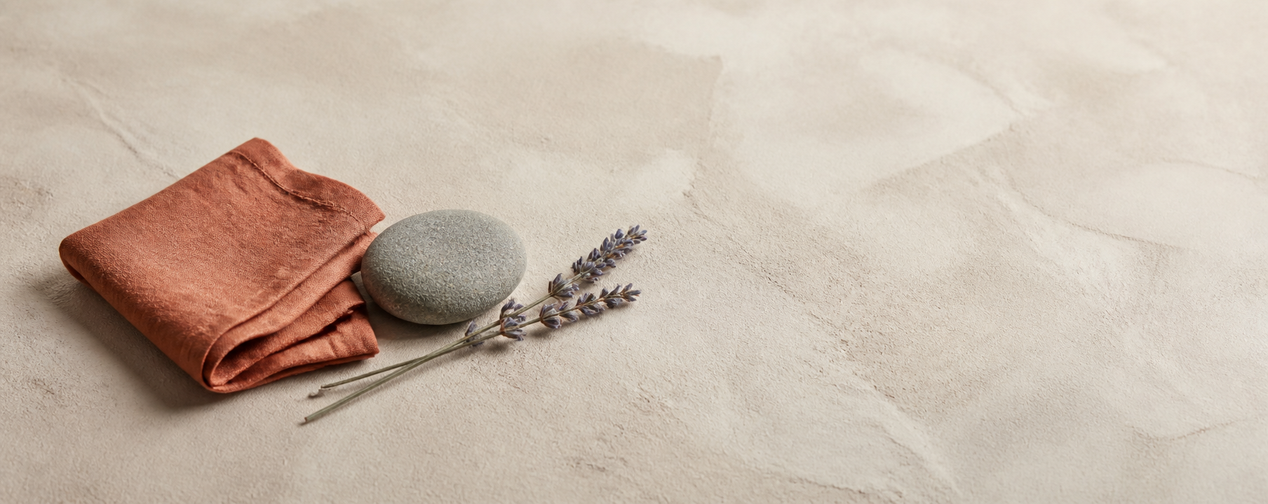 A folded terracotta linen cloth with a smooth gray stone and a dried sprig on pale sand