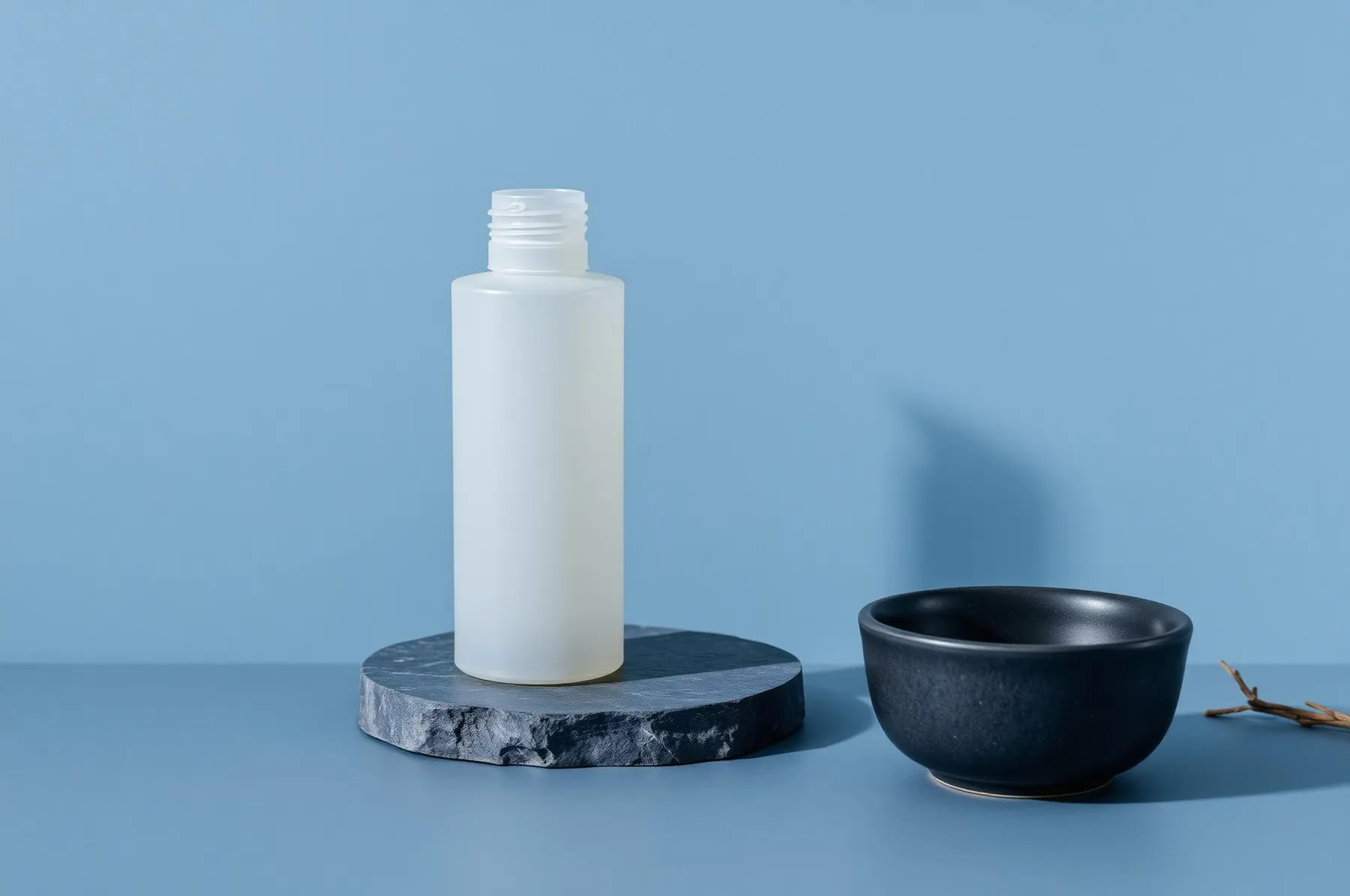 Tall unlabeled frosted bottle on a slate blue stone slab beside a matte charcoal dish