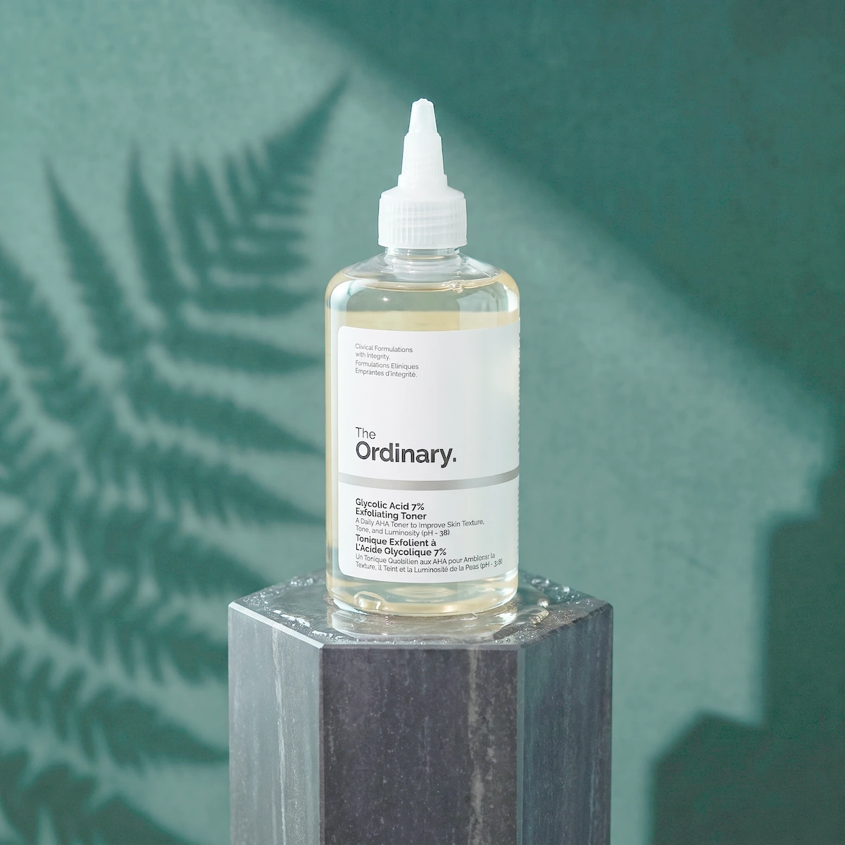 The Ordinary Glycolic Acid 7% Toning Solution bottle on wet dark stone, with a white cap and a leaf shadow