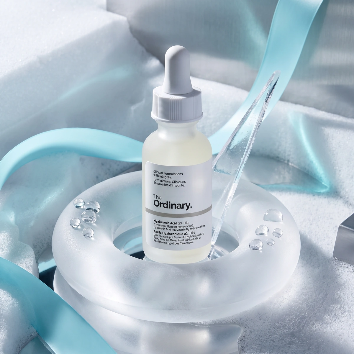 The Ordinary Hyaluronic Acid 2% + B5 dropper bottle on wet dark stone, with clear droplets and a leaf shadow